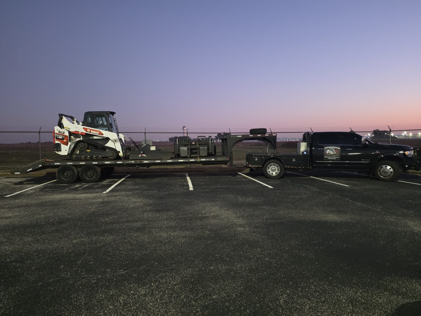 Black Ram dually flatbed pulling a gooseneck trailer loaded with a Bobcat T66 and attachments at sunrise